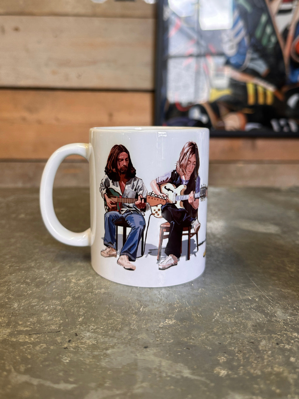 Beatles A white ceramic mug featuring a printed graphic of two men playing guitars, sitting on a rustic stone countertop with a wooden wall and framed art in the background.