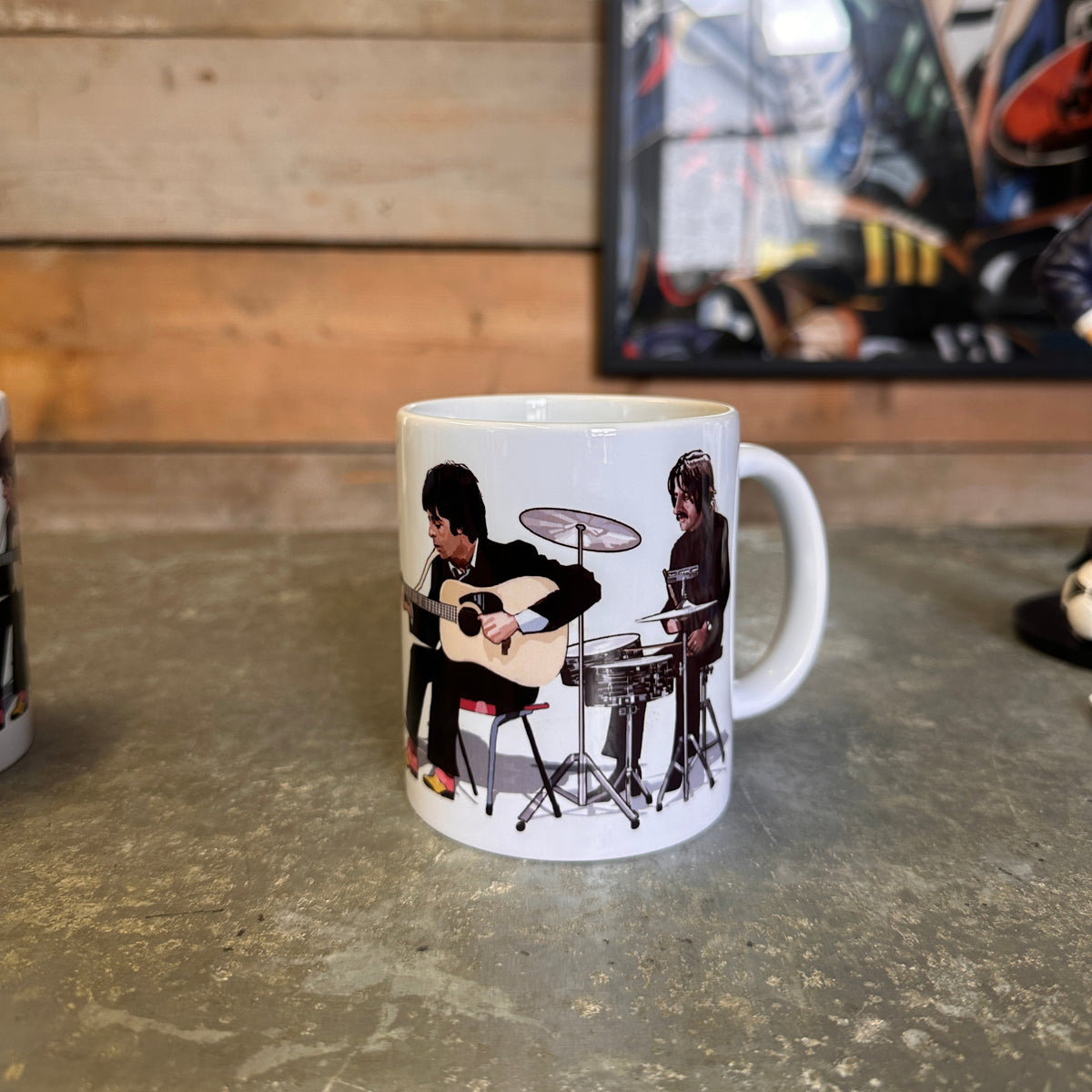 A white Beatles Mug  ceramic coffee mug featuring a printed graphic of two rock musicians, one playing an acoustic guitar and the other playing a drum set, sitting on a stone countertop against a wooden wall.